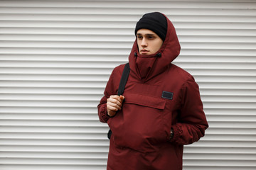 handsome model man in a warm fashionable jacket with a hood with a hat and a backpack near the metal wall on the street © alones