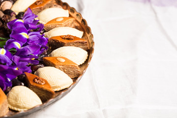 Fototapeta na wymiar Novruz copper tray with Azerbaijan national pastry pakhlava and shekerbura with purple fleur de lis flowers on white traditional kelagai silk female scarf background, spring new year celebration 