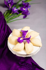 Novruz traditional pastry shekerbura in round white plate cake stand with bouquet purple lilac flowers fleur de lis on grey background, spring celebration in Azerbaijan, greeting card copy space