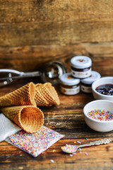 Fototapeta na wymiar A Kit For Making Ice Cream: Spoon For Ice Cream, Ice Cream Cones, Jam, Topping, Chocolate, Sprinkles, Coconut Shavings. On Wooden Background. The Concept Of A Set, Surprise, Gift Idea