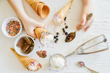 The child Makes his own Ice cream: Top View, Children's Hands Holding a cone of ice Cream On the table Chocolate, Spoon, Topping, Berries, Sprinkles, Jam.