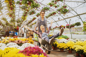 Two cheerful smiling florist women worker enjoying a break and ride into a cart in the greenhouse full of colourful flowers and pots.