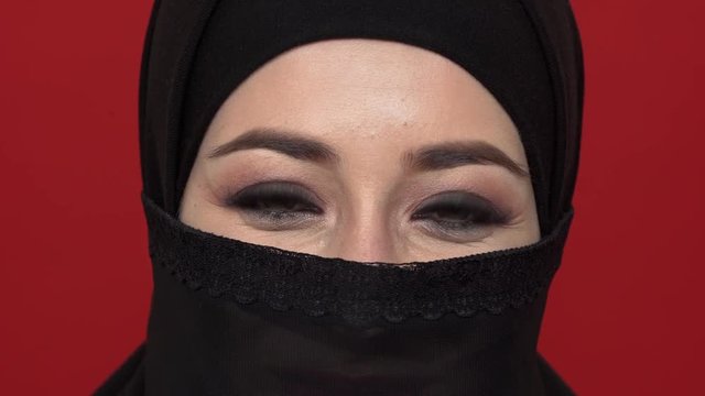 Macro portrait of middle eastern woman with smoky eyes visage in black niqab fooling around and crossing eyes, over red background. Concept of emotions