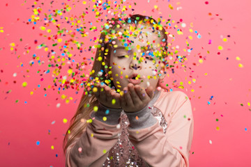 Happy girl celebrating on a pink background. Blows up multicolored confetti