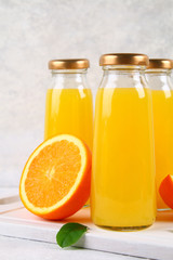 Glass bottles with fresh orange juice with orange slices and yellow tubes on a light gray table.