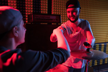 Portrait of tattooed modern singer fitstbumping drummer while rehearsing in dim recording studio