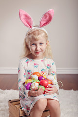 Cute adorable Caucasian child girl wearing Easter bunny rabbit ears sitting on wooden box in studio indoors. Kid holding holiday colorful eggs celebrating traditional holy Christian holiday