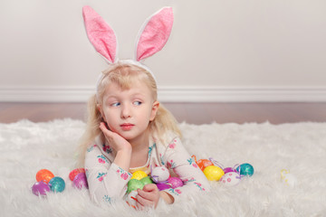 Cute adorable Caucasian child girl wearing Easter bunny rabbit ears lying on soft fluffy rug carpet in studio. Kid holding holiday colorful eggs celebrating traditional holy Christian holiday