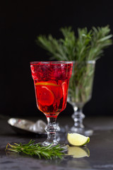 a glass with a cocktail of red color with lime, rosemary and ice cubes on a background of rosemary on a dark background