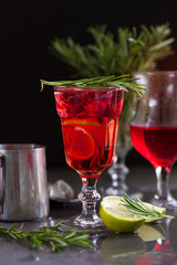 a glass with a cocktail of red color with lime, rosemary and ice cubes on a background of rosemary on a dark background