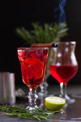 a glass with a cocktail of red color with lime, red berries, rosemary, burning cinnamon and ice cubes on a background of rosemary on a dark background