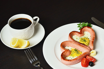 Scrambled eggs in the form of heart on a white plate with sausages, tomatoes, greens and coffee with lemon.