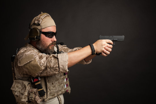 Brutal man in the military desert uniform and body armor standing in a fighting rack and aiming from his gun on a black background