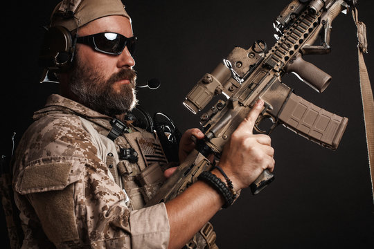 Brutal man in the military desert uniform and body armor stands in a fighting rack and holds his rifle on a black background