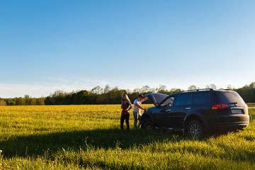 A young couple stands next to an off-road vehicle with an open hood. A girl calls to the technical service or a tow truck. The man bent over the engine while holding the hood with one hand.