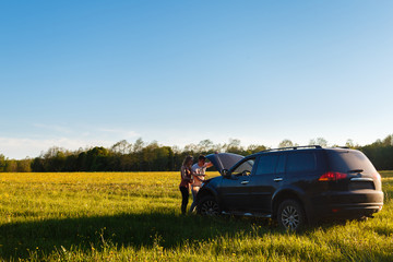 A young couple stands next to an off-road vehicle with an open hood. A man and a girl repair the engine of the car, holding the hood with one hand. The pair was stuck in the car on the field