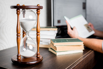 Hourglass or Sandglass at a desk showing time flow while a man reading books to learn the material with a computer on a background. Time concept. With a copy space
