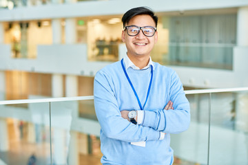Waist up portrait of cheerful Asian businessman wearing glasses smiling happily at camera standing at glass balcony in modern office building, copy space