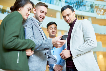 Obraz premium Low angle portrait of modern businessman holding smartphone showing something to group of colleagues standing in hall of office building