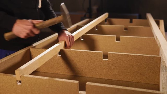 Joiner is installing detail of piece of furniture in a shop of factory. He is putting on a girder on a box and knocking by hammer