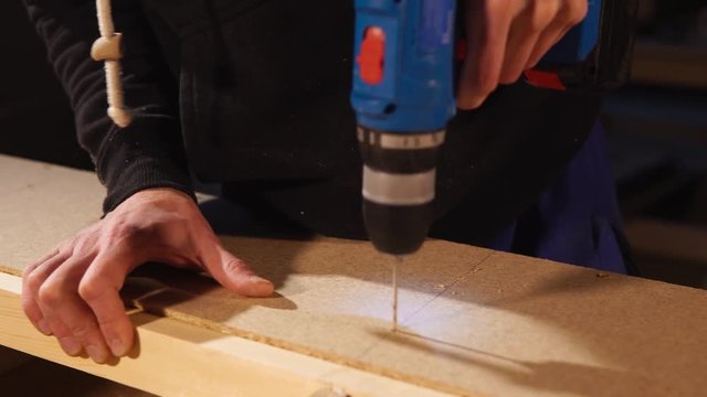 Joiner is assembling a furniture on a factory. He is using drill for making holes for fastening in a billet