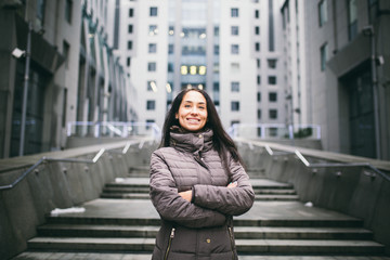 portrait of a woman on a business theme. Young caucasian brunette with long hair girl in long jacket, coat stands on business center background, office building with glass facade. Overcast in winter