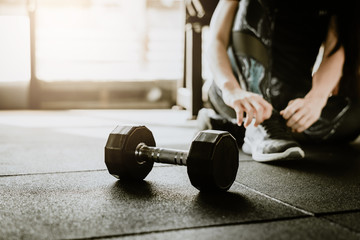 Dumbbell on the floor in gym with woman tying her shoelaces before weight trainning in background. fitness concept - obrazy, fototapety, plakaty