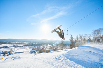 Naklejka na ściany i meble Full length action shot of young man performing snowboarding stunt spinning in air after backside flip in sunlight at ski resort, copy space