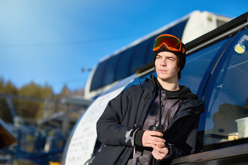Fototapeta na wymiar Portrait of modern young man wearing snowboarding gear relaxing leaning on truck and warming up with coffee, copy space