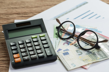 Tax calculation or budget planning concept, eyeglasses on pile of Euro banknotes on graph and chart performance print paper and calculator on wood table