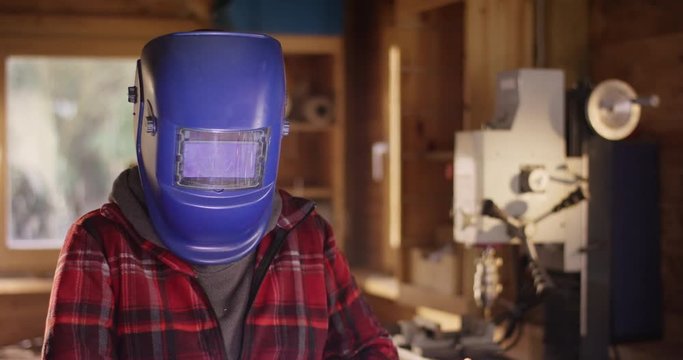 Young attractive woman welds metal in a workshop and reveals her beautiful face