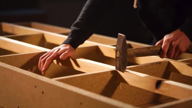 Close-up of assembling of furniture subject in a workshop of joinery. Master is knocking by hummer over wooden beam for installing