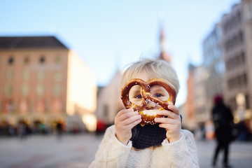 Little tourist holding traditional bavarian bread called pretzel on the town hall building background in Munich, Germany - obrazy, fototapety, plakaty