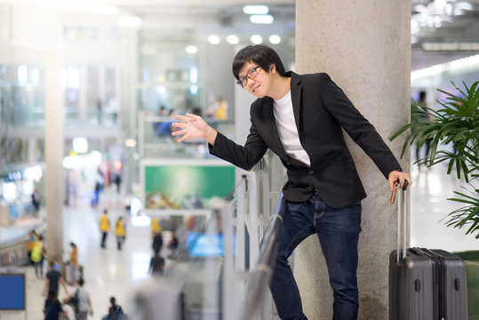 Young asian man with suitcase luggage raising hand for greeting with his friend in the international airport terminal, business travel concept