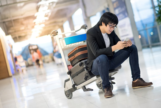 Young Asian man using smartphone sitting on airport trolley with his suitcase luggage in the international airport terminal, business travel concepts