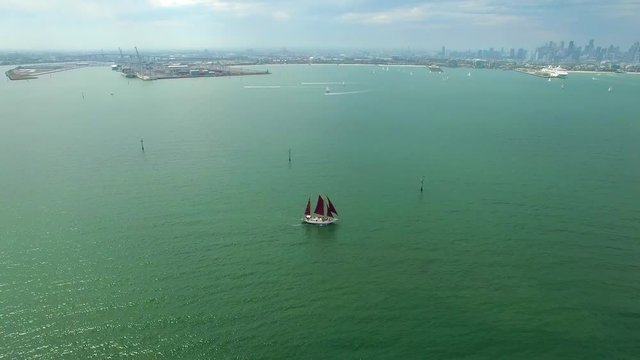 Backward flight away from beautiful sailboat with dark red sails sailing across ocean bay