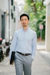 A portrait of a young Chinese Asian businessman walking down a street in Asia. He is dressed in a professional blue shirt and pants for the day. He is smiling and carrying his leather folio with him.