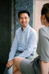 A young Chinese man who is professionally dressed is taking a break and having a discussion with his colleague in their office. He is smiling and is relaxed and natural. 