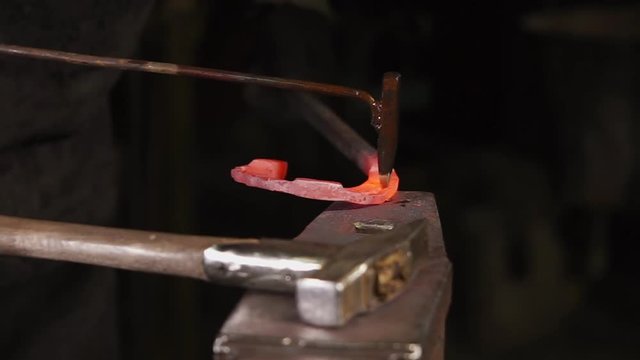 Steel horseshoe is lying over large incus. Blacksmiths are beating on it using chisel, hammer and maul