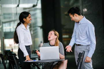 A diverse group of professionals are sitting around a table and engaging in a business plan and having a discussion. As a group, a Chinese man, a Caucasian woman and an Indian woman speak.