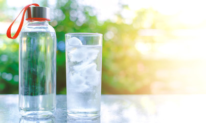 Water bottle and a glass of ice on blurred natural green background with copy space and sun light effect for healthy drinking concept