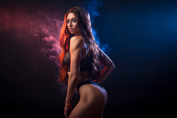 Young dark-haired woman in black lingerie posing against a background of red and   blue smoke from a vape on a black isolated background