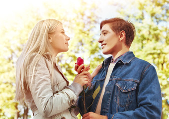 Spring couple walking and kissing summer park. Boyfriend and girlfriend walk in sun shine. Guy gives girl red rose flower. Fairy-tale love at first sight.