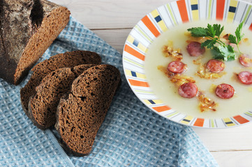 Soup puree from potatoes with smoked sausages and fried onions. The dish is decorated with a sprig of parsley. In the frame, cloth napkin and a few slices of bread. Wooden light background. Close-up.