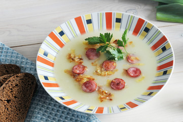 Soup puree from potatoes with smoked sausages and fried onions. The dish is decorated with a sprig of parsley. In the frame, cloth napkin and a few slices of bread. Wooden light background. Close-up.
