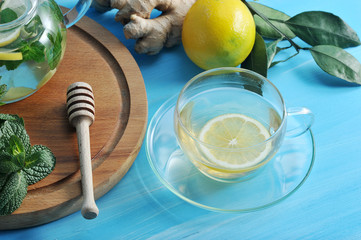 A cup with a warming ginger drink. In the frame, the root of ginger, a bunch of mint, lemon. Blue wood background. Close-up.
