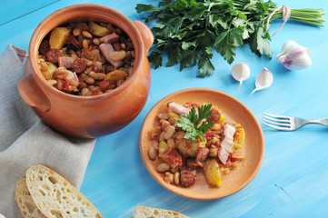 Beans in a rustic style with sausages in earthenware. Next a plate with cooked beans and a fork. A bunch of parsley, white bread, garlic complement the composition. Blue background. Close-up.