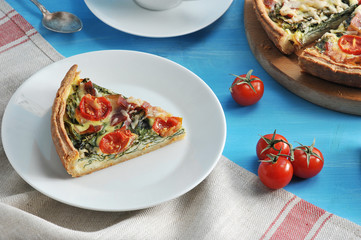 A piece of pie quiche with spinach, bacon, cherry tomatoes and cheese. In the frame, a piece of cake on a platter and cherry tomatoes. Blue background draped with a napkin. Close-up.