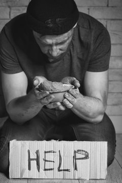 Homeless poor man holding carton board with word HELP and bowl for donations while sitting near brick wall