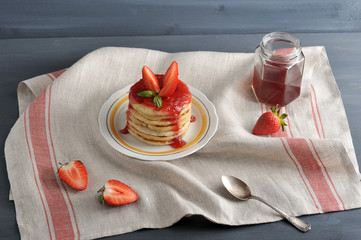 On a plate of pancakes with strawberry jam, decorated with fresh strawberries. Next to the bank with strawberry jam, a teaspoon, a strawberry. The background is draped with a napkin. Close-up.
